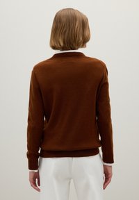 Pull en tricot marron avec un col et des poignets côtelés, présentant une coupe décontractée et un tissu texturé, associé à un pantalon blanc.