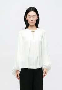 Blouse blanche à manches longues avec un col en V, un devant plissé et un détail en métal doré au niveau de l'encolure. Tissu lisse et fluide avec des poignets froncés.