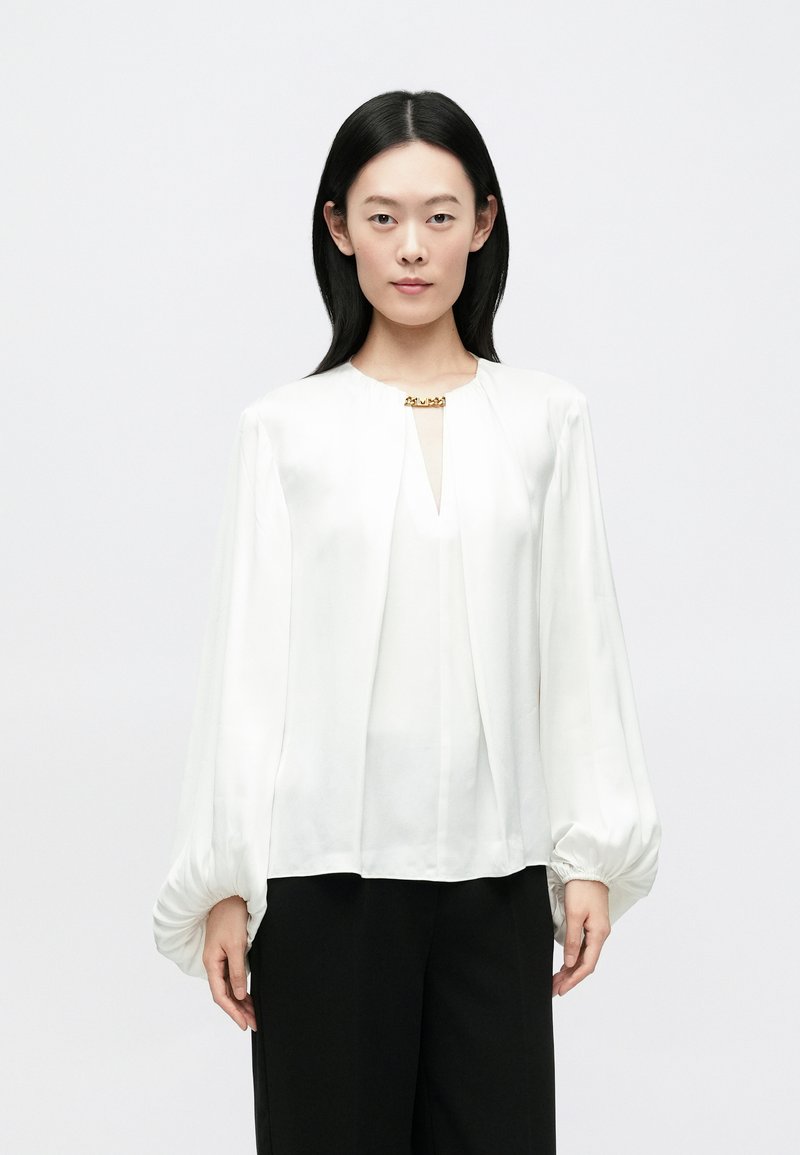 Blouse blanche à manches longues avec un col en V, un devant plissé et un détail en métal doré au niveau de l'encolure. Tissu lisse et fluide avec des poignets froncés.