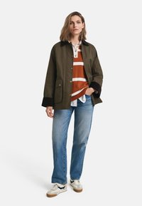Green button-up jacket with chest pockets, brown collar, layered over an orange-striped sweater and blue jeans. White sneakers complete the look.