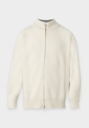 Homme debout portant un pull côtelé crème à fermeture éclair, un pantalon sombre et des baskets marron, une main dans la poche, sur un fond uni.