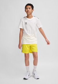 Weißes Baumwoll-T-Shirt mit schwarzem Schulterbesatz und Logo, kombiniert mit leuchtend gelben Shorts mit seitlichen Akzenten. Weiße Sportschuhe und Socken.