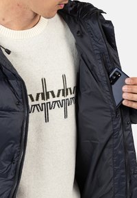 Giacca isolante blu navy con tasca con zip, indossata sopra un maglione crema con un motivo geometrico in nero e verde. Smartphone visibile nella tasca.