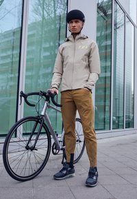 Beige jacka med reflekterande "FOX"-logotyp, bruna byxor, svart hjälm och grå cykel med svarta hjul. Stadsmiljö med glasbyggnader i bakgrunden.