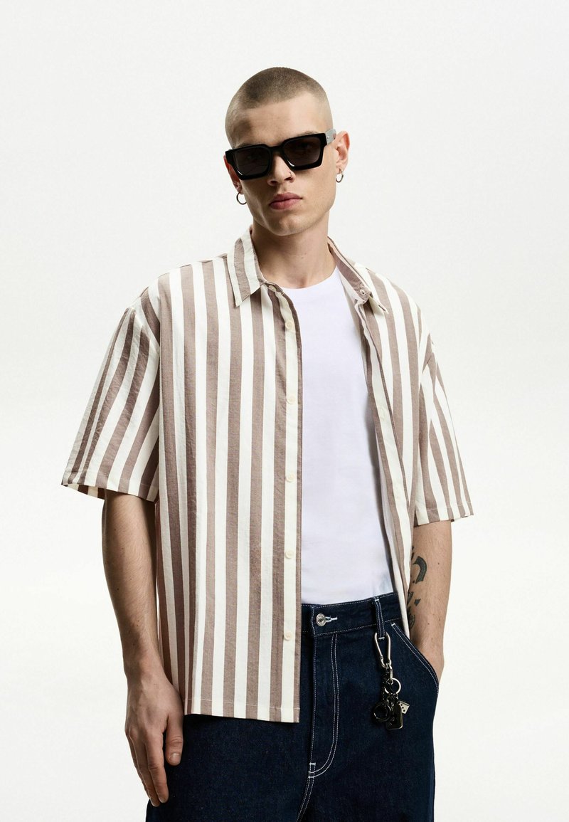 Short-sleeve button-up shirt in beige and white vertical stripes, paired with a plain white t-shirt and dark denim pants featuring a hardware accent.