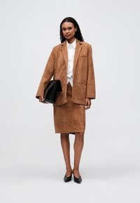 Blazer in suede marrone sopra una camicia bianca e una gonna in suede marrone abbinata. Clutch nera e tacchi neri. Design semplice senza motivi visibili.