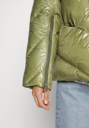 Manche de veste matelassée verte avec détail de fermeture éclair visible sur le bras, main détendue sur le côté, associée à un jean bleu clair.