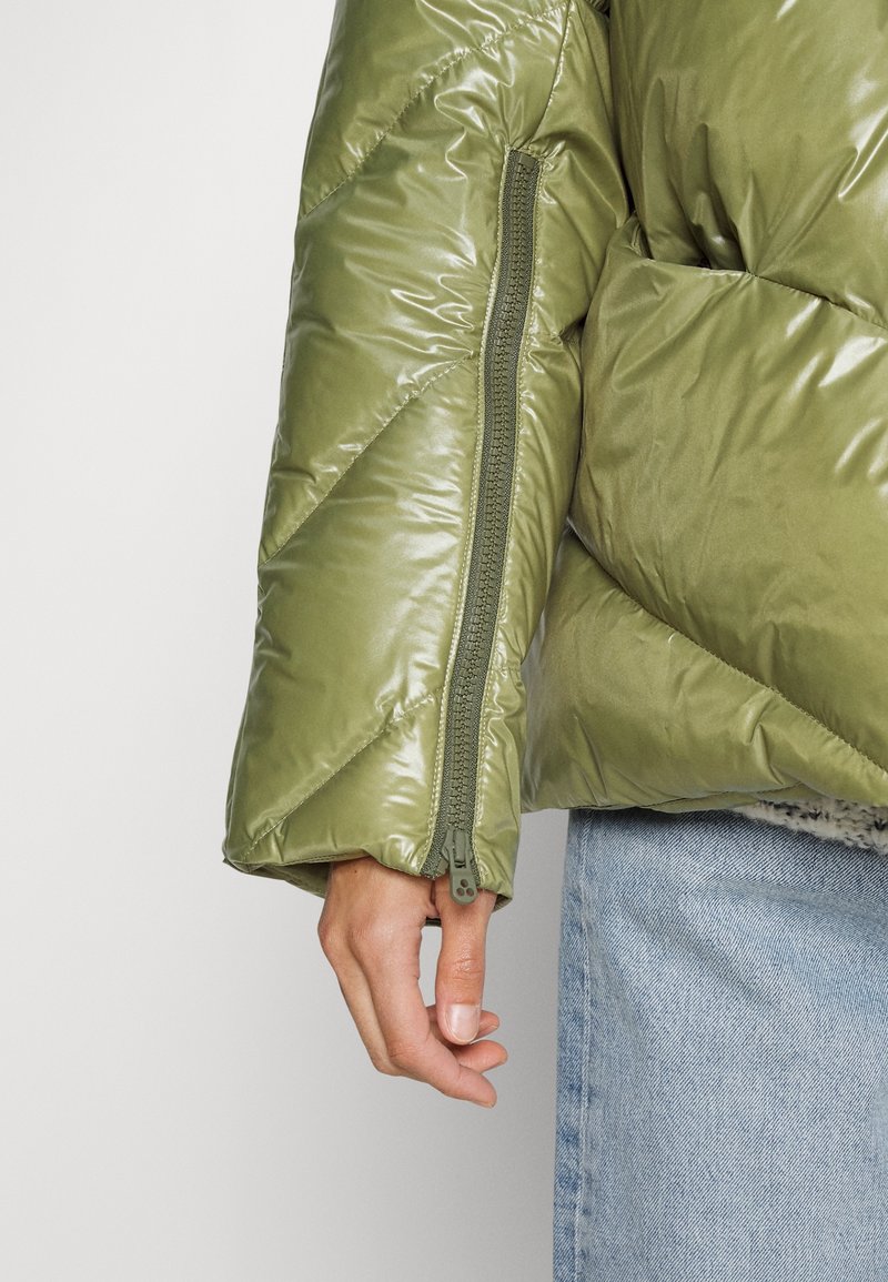 Manche de veste matelassée verte avec détail de fermeture éclair visible sur le bras, main détendue sur le côté, associée à un jean bleu clair.