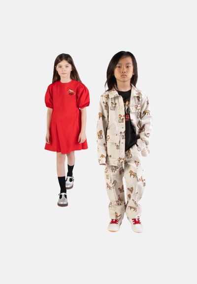 Twee kinderen staan, de een draagt een rode jurk met opgeblazen mouwen, de ander een beige jas met paardenprint en een bijpassende broek, met rood gestrikte schoenen.