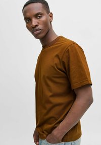 Camiseta de algodón marrón con un clásico cuello redondo y mangas cortas, mostrada ligeramente descentrada sobre un fondo claro.