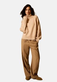 Maglione beige a collo alto in maglia a coste con maniche larghe, abbinato a pantaloni beige a gamba larga con cuciture verticali.