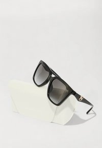 Ochelari de soare oversized negri cu lentile în degrade, cadru din plastic, accente din metal auriu pe braț, așezați pe un toc alb cu logo embossat.