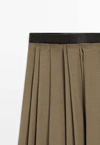Jupe plissée vert olive taille haute avec une large ceinture noire, montrant des plis structurés et une texture de tissu lisse.