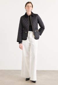 Gequilted zwarte jas met ritssluiting, platte kraag en twee voorkuiten; gecombineerd met brede witte broeken en zwarte puntige hakken.