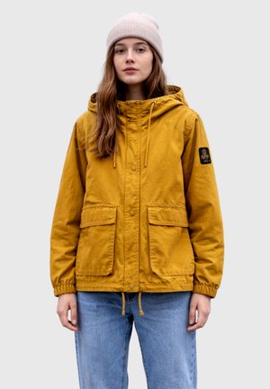 Giovane donna che indossa una giacca con cappuccio giallo senape con grandi tasche frontali, jeans azzurri chiari e un berretto lavorato a maglia beige, in piedi su sfondo grigio.