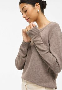 Pull en tricot marron avec des poignets côtelés et une coupe décontractée, présentant un col rond et une texture douce. Le modèle croise délicatement les mains.