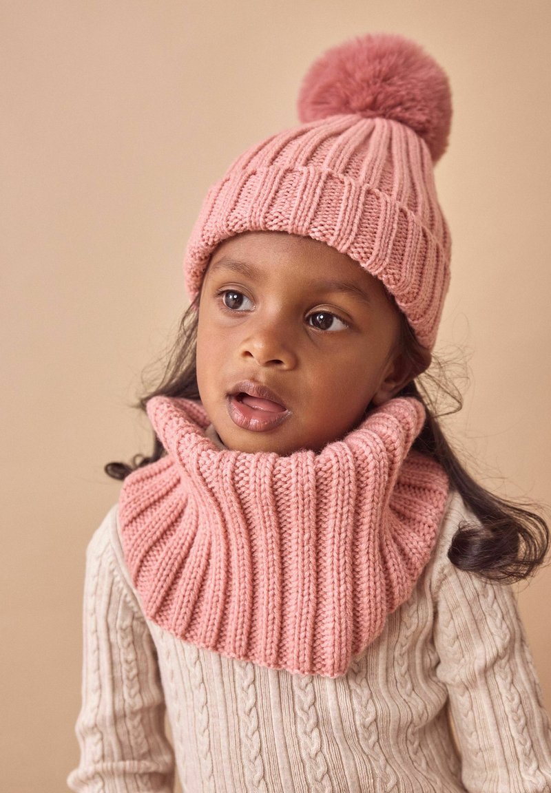 Rosa gestrickte Mütze mit einem Pompon und passendem gerippten Halswärmer. Der Pullover hat ein hellbeiges Zopfmuster. Glatte Textur.