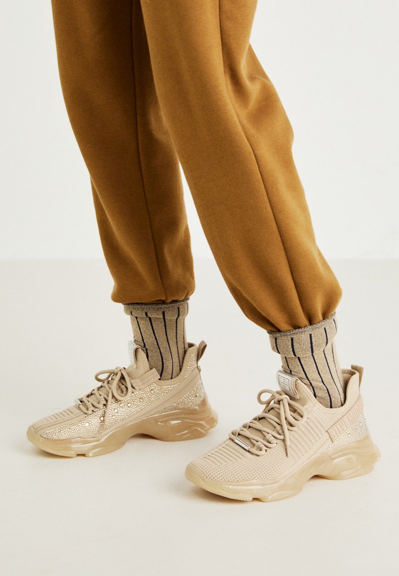 Beige athletic shoes with mesh upper, decorative sparkles, and thick rubber sole. Paired with striped beige socks and brown jogger pants.