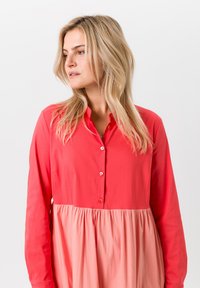 Koraalkleurige blousejurk met een aansluitende bovenkant, lange mouwen en een vloeiende rok in een lichtere tint, gemaakt van lichtgewicht stof.