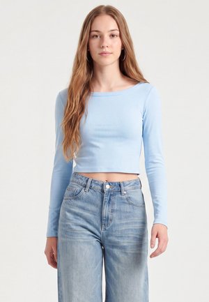 Ung kvinde med langt lysebrunt hår iført en lyseblå crop top med lange ærmer og højtsiddende, sløvede blå jeans, der står mod en ensfarvet baggrund.