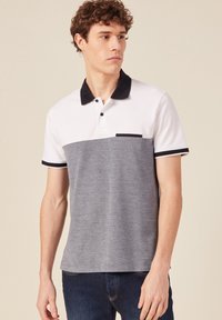 Camisa polo para hombre con diseño en bloques de color blanco y gris, mangas cortas, cuello negro y un pequeño bolsillo en el pecho.