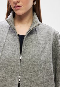 Γκρί ζακέτα από μίγμα μαλλιού με ψηλό γιακά, κλείσιμο με φερμουάρ και ριμπελίκες λεπτομέρειες. Ξεχωριστό ασημί υλικό και μαλακή υφή σε όλη την επιφάνεια.