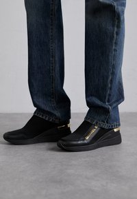 Sorte slip-on sneakers med mesh-stof, guld-detaljer og branding; parret med mørke denimjeans. Har et slankt, moderne design.