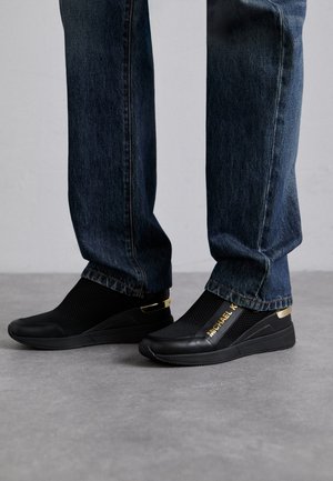 Sneakers neri slip-on in tessuto a rete, con dettagli dorati e branding; abbinati a jeans di denim scuri. Presentano un design elegante e moderno.