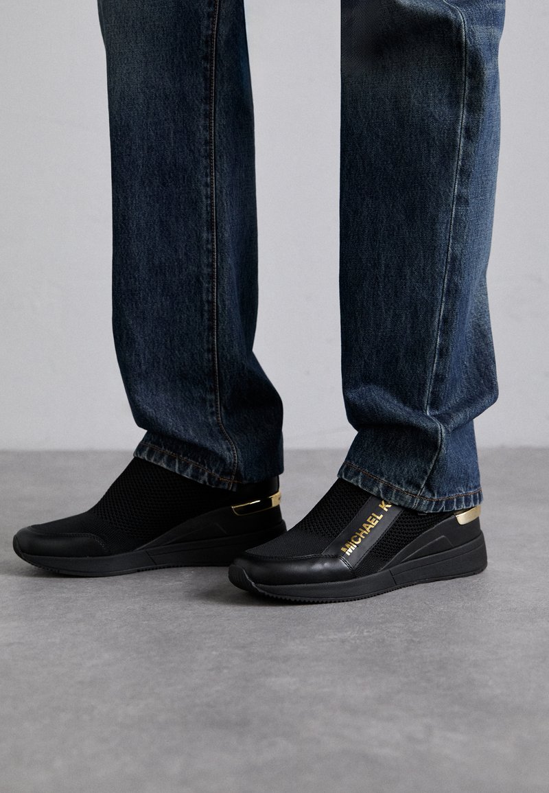Sorte slip-on sneakers med mesh-stof, guld-detaljer og branding; parret med mørke denimjeans. Har et slankt, moderne design.