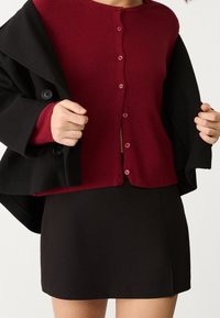 Maroon ribbed cardigan with buttons, paired with a black coat shown partially open and a black skirt. Smooth fabric and simple design.