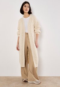 Langer beiger Strickcardigan mit Kabelmuster, offenem Vorderteil und weiten Ärmeln, getragen über einem weißen T-Shirt und beigen weiten Hosen.
