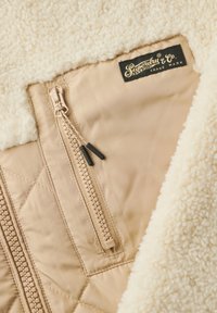 Tissu matelassé beige avec un col en fausse laine crème, équipé d'une fermeture éclair contrastante et d'une étiquette noire "Superdry" sur la couture.