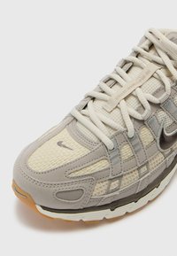 Buty sportowe z jasnoszarym i kremowym siateczkowym wierzchem z teksturowanymi detalami, sznurowadłami oraz gumową podeszwą. Zawiera srebrne akcenty i branding.