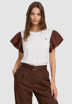 Donna che indossa una maglietta bianca con maniche corte marroni arricciate e pantaloni marroni a vita alta con cintura, in piedi con una mano in tasca.