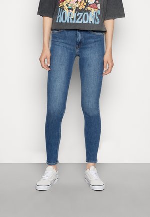 Personne portant un jean skinny bleu, des baskets blanches et un T-shirt noir avec des champignons et le mot "HORIZONS", debout devant un fond blanc.