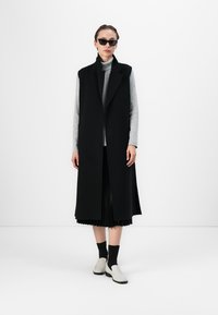 Garais melnais vestes ar apkakli, valkāts virs pelēka tārtnīša un piešūtā melnā svārka. Modelis nēsā melnus saulesbrilles un gaišas kurpes.
