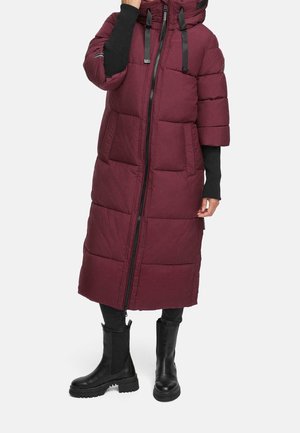 Personne portant un long manteau matelassé bordeaux avec une fermeture éclair à l'avant, un pantalon noir et des bottes noires épaisses sur un fond uni.