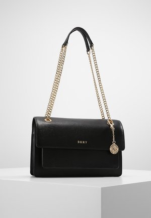 Sac à bandoulière en cuir noir DKNY avec des bretelles en chaîne dorée et un pendentif logo rond doré, posé sur un socle blanc sur un fond uni.