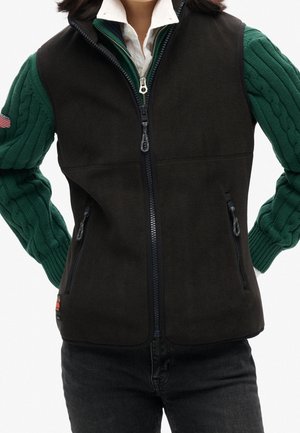 Persoon die een zwart fleecevest met rits draagt over een groene kabelgebreide trui en een wit overhemd met kraag, handen rustend op de heupen.