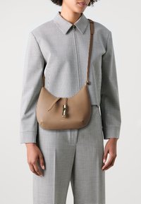Borsa in pelle marrone dalla forma curvata, caratterizzata da una cerniera centrale con accessori in oro lucido, indossata sopra una camicia a scacchi e pantaloni.