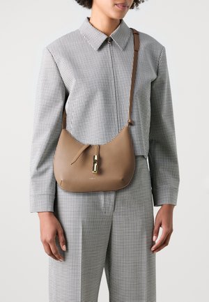 Borsa in pelle marrone dalla forma curvata, caratterizzata da una cerniera centrale con accessori in oro lucido, indossata sopra una camicia a scacchi e pantaloni.