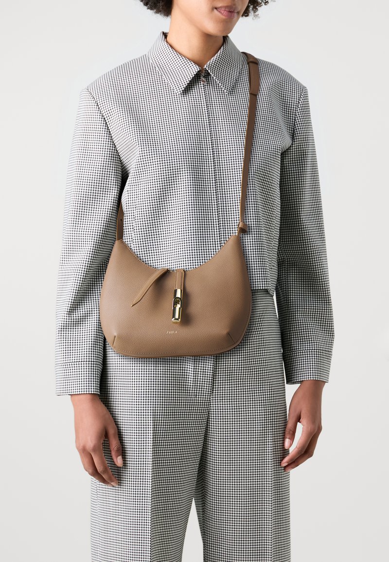 Borsa in pelle marrone dalla forma curvata, caratterizzata da una cerniera centrale con accessori in oro lucido, indossata sopra una camicia a scacchi e pantaloni.