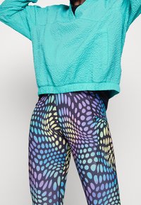 Top corto turchese texturizzato con una vestibilità comoda abbinato a leggings multicolori con un motivo a pois dinamico su uno sfondo scuro.