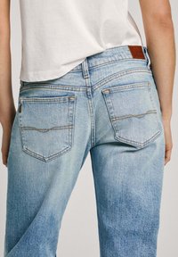 Los jeans de mezclilla azul claro cuentan con bolsillos traseros y un parche de cuero marrón en la cintura. La tela tiene un acabado desgastado con costuras visibles.
