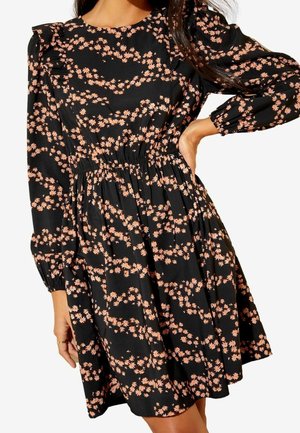 Femme portant une robe noire à manches longues avec un motif floral pêche, avec une taille cintrée et des épaules légèrement bouffantes.