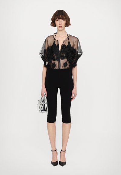 Schwarze, transparente Bluse mit Blumenmuster und kurzen Ärmeln, kombiniert mit knielangen schwarzen Leggings und schwarzen spitzen Heels, während sie eine kleine silberne Tasche hält.