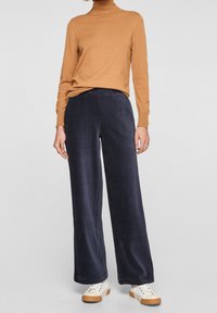 Hnědý proužkovaný rolák spárovaný s námořnickými širokými kalhotami z kordu. Tenisky mají krémový svršek a gumovou podrážku. Neutrální pozadí.