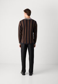 Uomo di spalle con un maglione a righe verticali marroni e nere, pantaloni neri e scarpe nere su uno sfondo bianco puro.