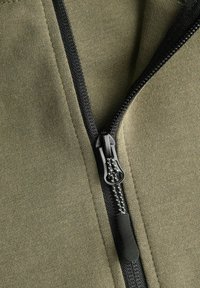 Tissu vert olive avec une texture lisse, comportant une fermeture éclair noire traversant le centre et un détail de tirette texturée.