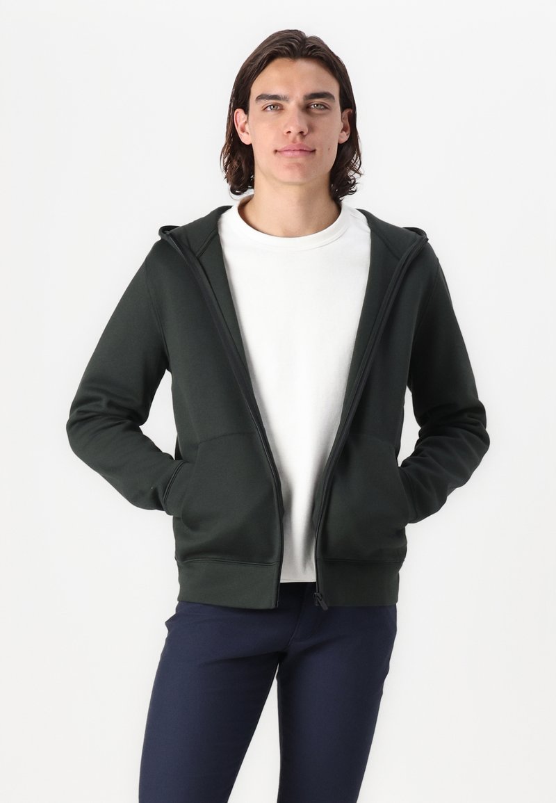 Ciemnozielona bluza z kapturem na zamek błyskawiczny, wykonana z miękkiego materiału. Posiada kieszenie z przodu i elastyczne mankiety. Noszona na białej koszulce, w komplecie z granatowymi spodniami.